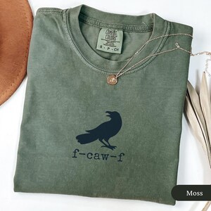 May include: A moss green t-shirt with a black raven graphic and the text "f-caw-f". A gold-colored necklace with a pendant rests on the shirt. The shirt is made of a soft material.