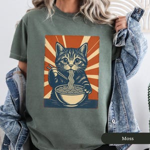 May include: A moss green t-shirt featuring a graphic of a cat eating ramen noodles. The cat is depicted in a vintage style with a bowl of noodles and chopsticks. The background has a retro sunburst design in orange and cream.