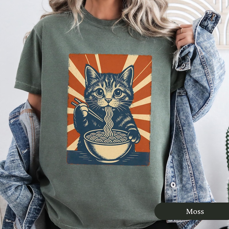 May include: A moss green t-shirt featuring a graphic of a cat eating ramen noodles. The cat is depicted in a vintage style with a bowl of noodles and chopsticks. The background has a retro sunburst design in orange and cream.