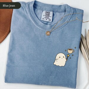 May include: A blue Comfort Colors t-shirt with a ghost graphic holding a magic wand and a coffee cup. A gold necklace with a pendant rests on the shirt. The shirt is labeled "Blue Jean".