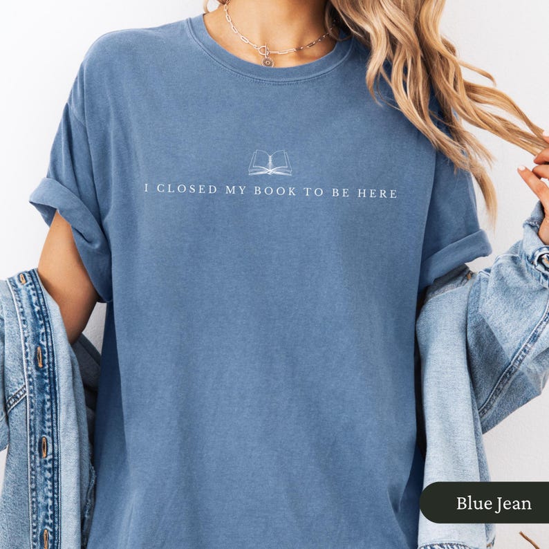 May include: Blue jean-colored t-shirt with the text "I CLOSED MY BOOK TO BE HERE" and a small book graphic. The shirt has rolled-up sleeves and a relaxed fit. The color is a faded denim blue.