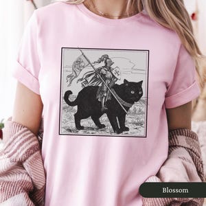 May include: A pink t-shirt with a black and white illustration of a knight riding a black cat. The knight is holding a spear and a shield. The illustration is in a square frame.