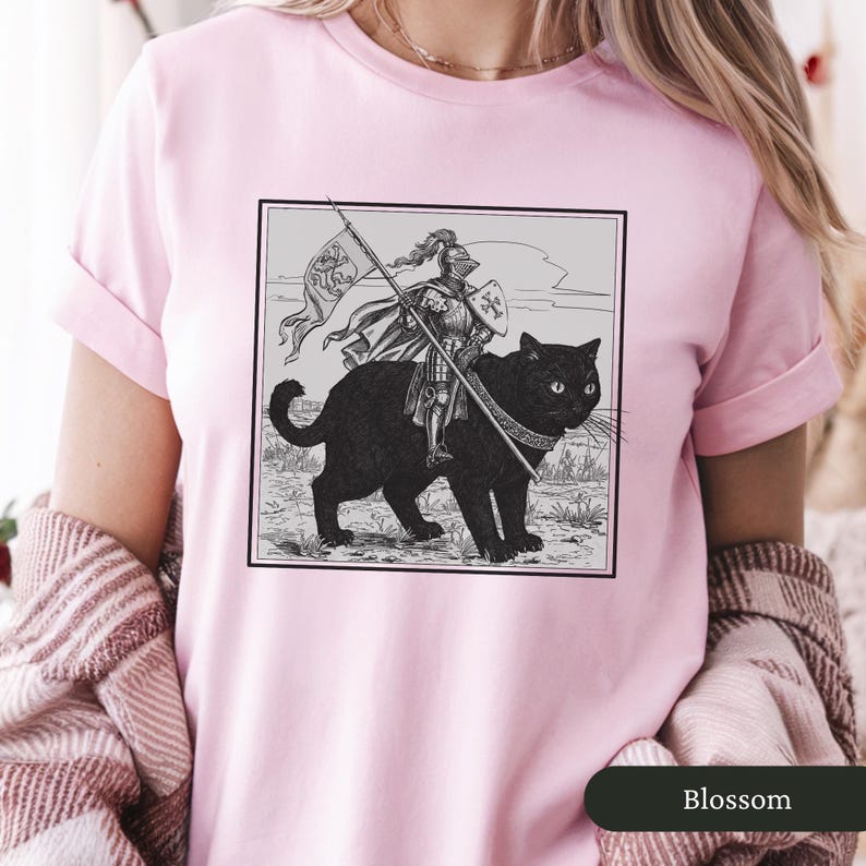May include: A pink t-shirt with a black and white illustration of a knight riding a black cat. The knight is holding a spear and a shield. The illustration is in a square frame.