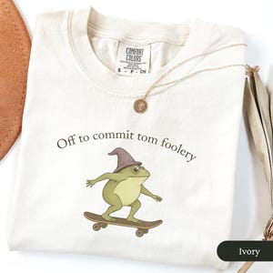 Puede incluir: Camiseta de color marfil con la frase "Off to commit tom foolery" y una rana de dibujos animados con un sombrero de mago y montada en monopat&iacute;n. Tambi&eacute;n se ve un collar de oro con un colgante.