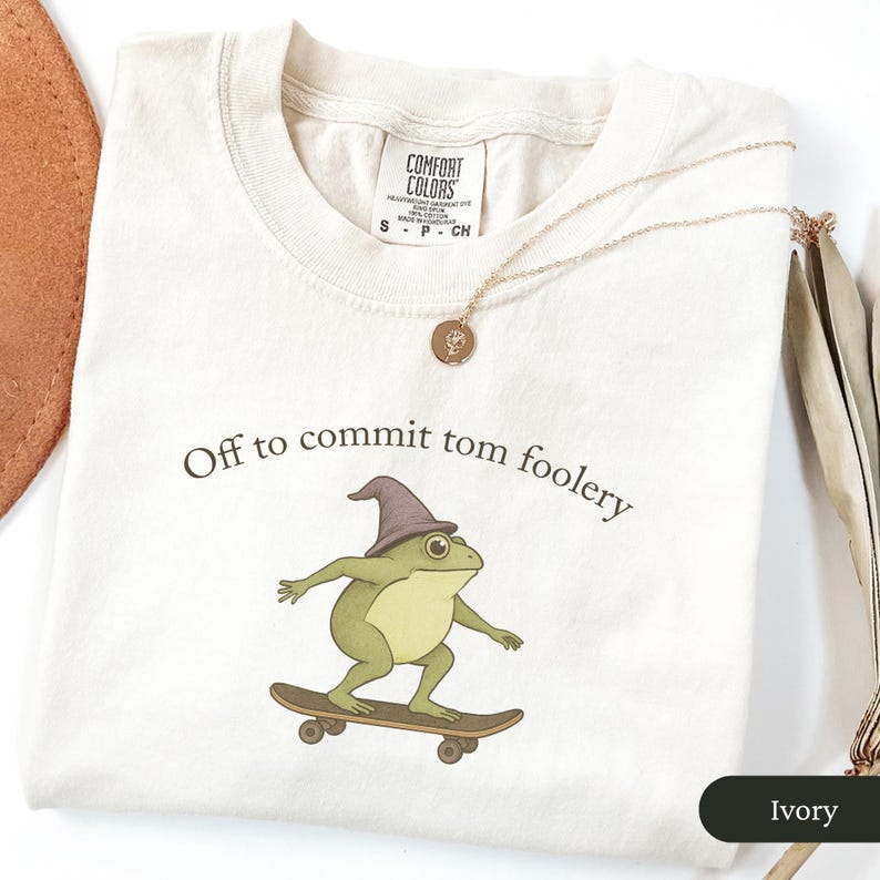 Puede incluir: Camiseta de color marfil con la frase "Off to commit tom foolery" y una rana de dibujos animados con un sombrero de mago y montada en monopat&iacute;n. Tambi&eacute;n se ve un collar de oro con un colgante.