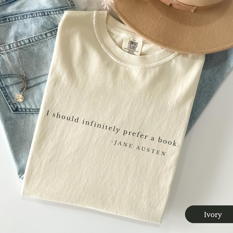 Puede incluir: Camiseta color marfil con la cita "I should infinitely prefer a book." de Jane Austen. La camiseta est&aacute; doblada sobre unos vaqueros azules, con un collar y un sombrero beige en el fondo.