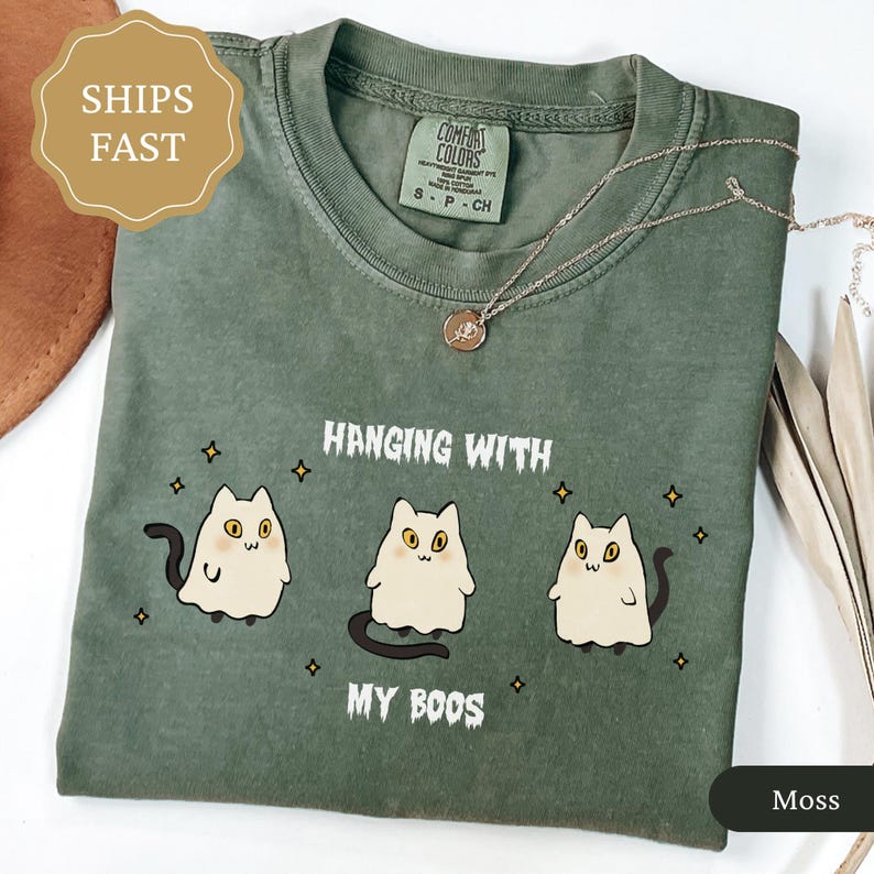 May include: A moss green t-shirt featuring the text "Hanging with my Boos" and three ghost cat illustrations. The shirt has a crew neck and short sleeves. A gold necklace with a pendant is on the shirt.