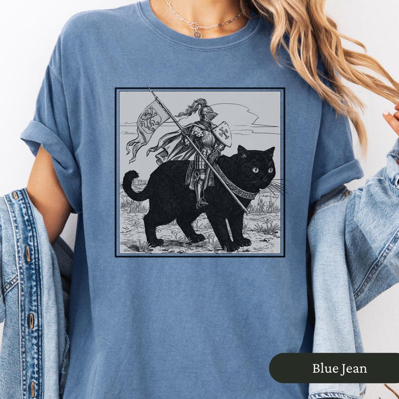 May include: A blue jean t-shirt with a black and white illustration of a knight riding a black cat. The knight is holding a spear and a flag with a cross on it. The illustration is in a square frame.