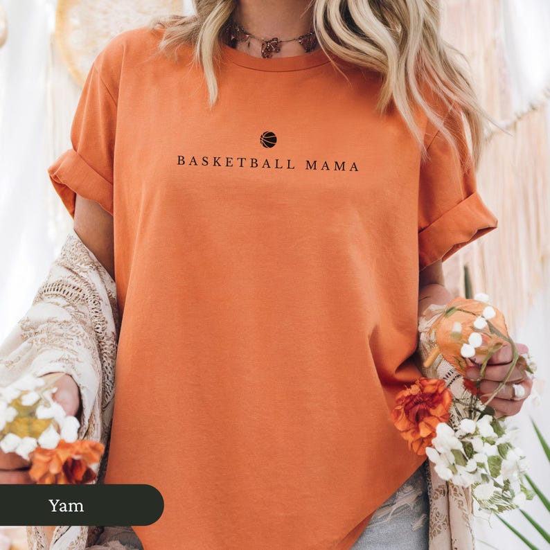 以下が含まれることがあります： バスケットボールのグラフィックの上に「BASKETBALL MAMA」の文字が入った、袖をまくったオレンジ色のTシャツ。柔らかい素材で作られたシャツは、カジュアルウェアです。
