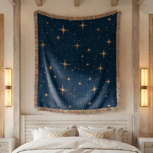 Puede incluir: Una manta tejida azul marino con un estampado de estrellas celestiales. La manta presenta estrellas doradas de varios tama&ntilde;os y un borde con flecos. Se muestra sobre una cama con ropa de cama blanca.