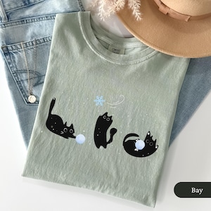 Puede incluir: Una camiseta verde claro con un dise&ntilde;o l&uacute;dico de tres gatos negros interactuando con una bola azul y un copo de nieve. La camiseta est&aacute; doblada sobre unos vaqueros azules y un sombrero.