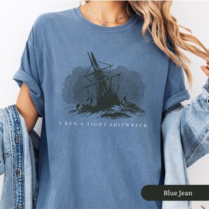 May include: Blue jean-colored t-shirt featuring a distressed graphic of a shipwreck. The text "I RUN A TIGHT SHIPWRECK" is printed below the image. The shirt has rolled-up sleeves and a relaxed fit.
