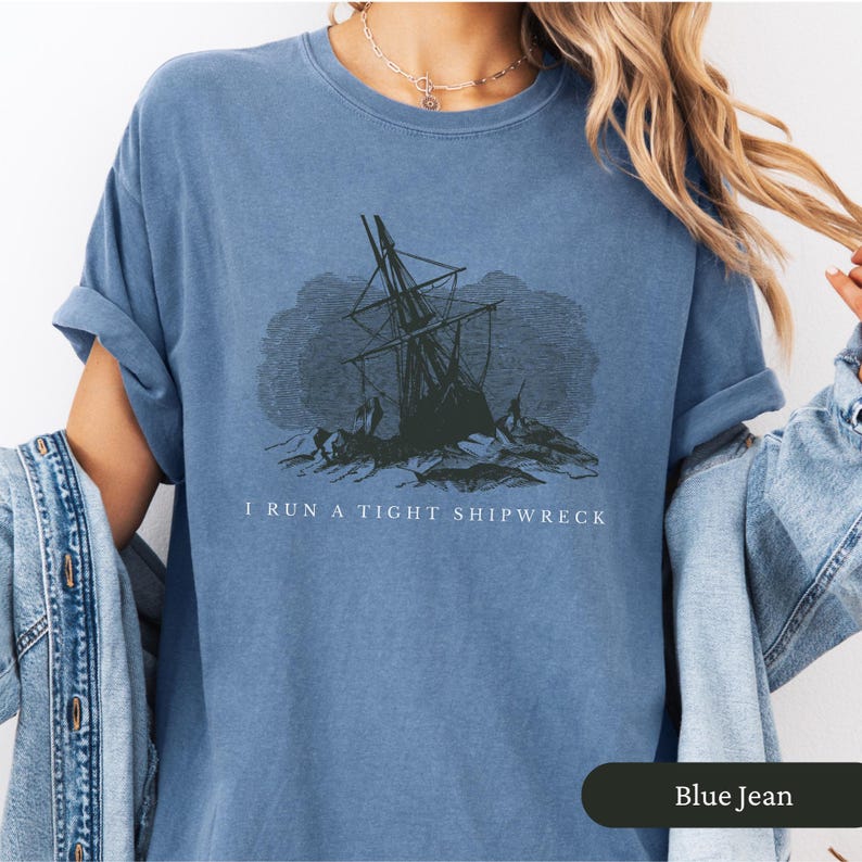 May include: Blue jean-colored t-shirt featuring a distressed graphic of a shipwreck. The text "I RUN A TIGHT SHIPWRECK" is printed below the image. The shirt has rolled-up sleeves and a relaxed fit.