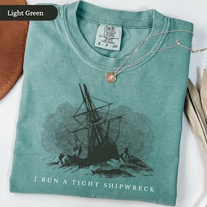 May include: Light green Comfort Colors t-shirt with a distressed graphic of a shipwreck and the text "I RUN A TIGHT SHIPWRECK." A silver necklace with a small pendant rests on the shirt. The shirt is folded and displayed on a white surface.