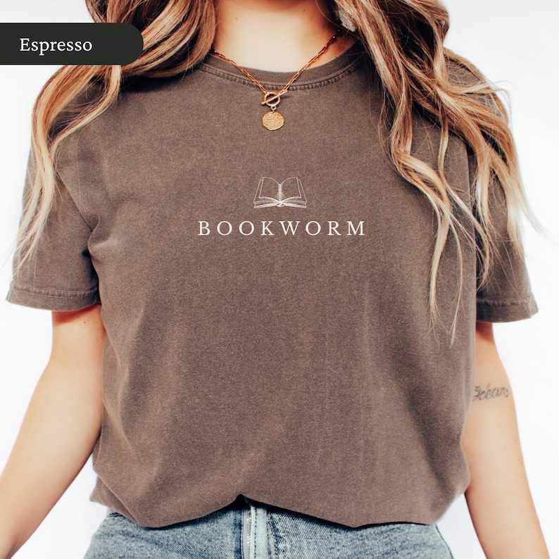Op de afbeelding: Espresso-kleurig T-shirt met het woord "BOOKWORM" in het wit gedrukt onder een lijntekening van een open boek. Het shirt heeft korte mouwen en een ronde hals.