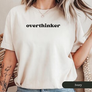 May include: Ivory-coloured t-shirt featuring the word "overthinker" in black. The shirt has short sleeves and a crew neck. The image shows a close-up of the shirt, highlighting the text and fabric texture. The shirt is a neutral colour.