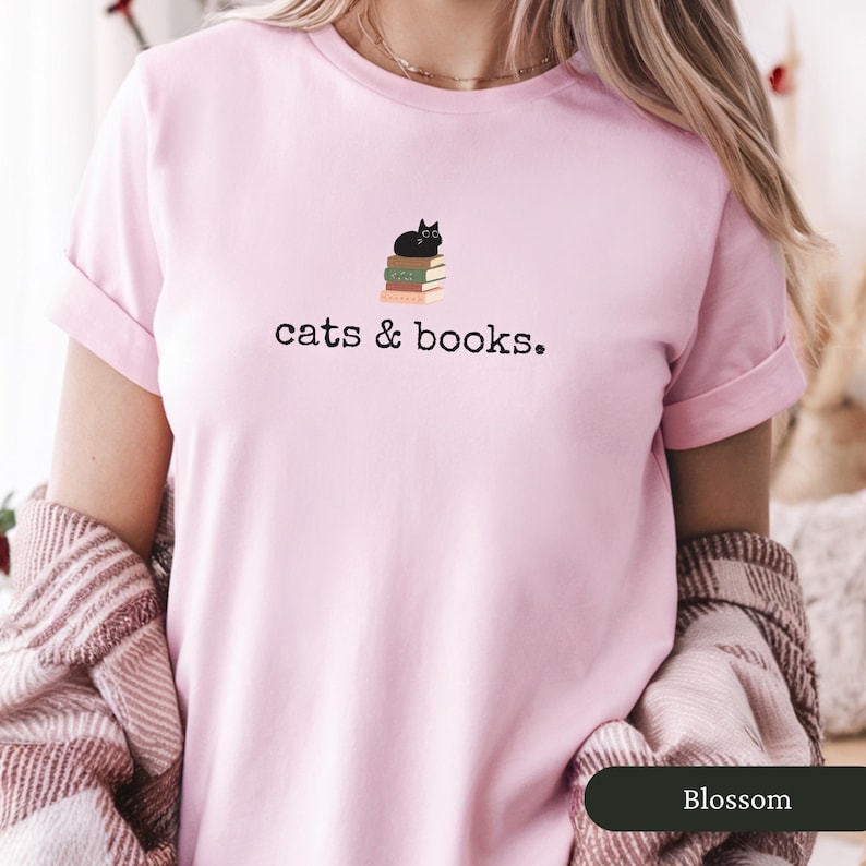 Może przedstawiać: R&oacute;żowa koszulka z czarnym kotem siedzącym na stosie książek. Na koszulce nadrukowany jest napis "cats & books."