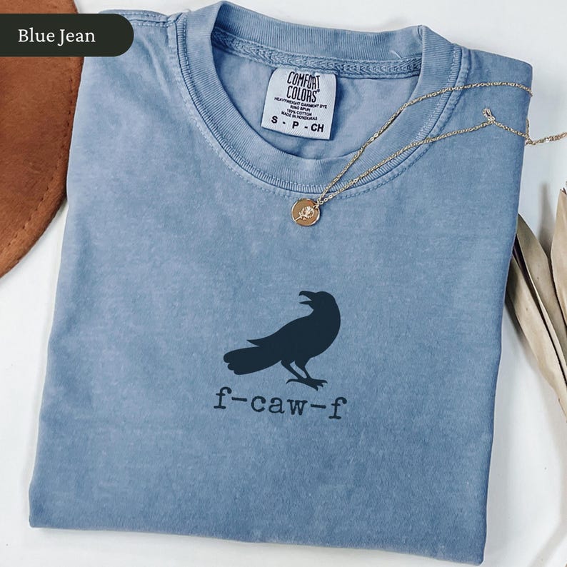 May include: Blue jean colored t-shirt with a black crow graphic and the text "f-caw-f". The shirt has a crew neck and short sleeves. A gold necklace with a small pendant is draped across the shirt.