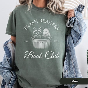 Puede incluir: Una camiseta verde con un dise&ntilde;o gr&aacute;fico blanco que dice "Trash Readers Book Club" con un mapache y una rata leyendo libros en un contenedor de basura. El texto tambi&eacute;n dice "Est. 2025".