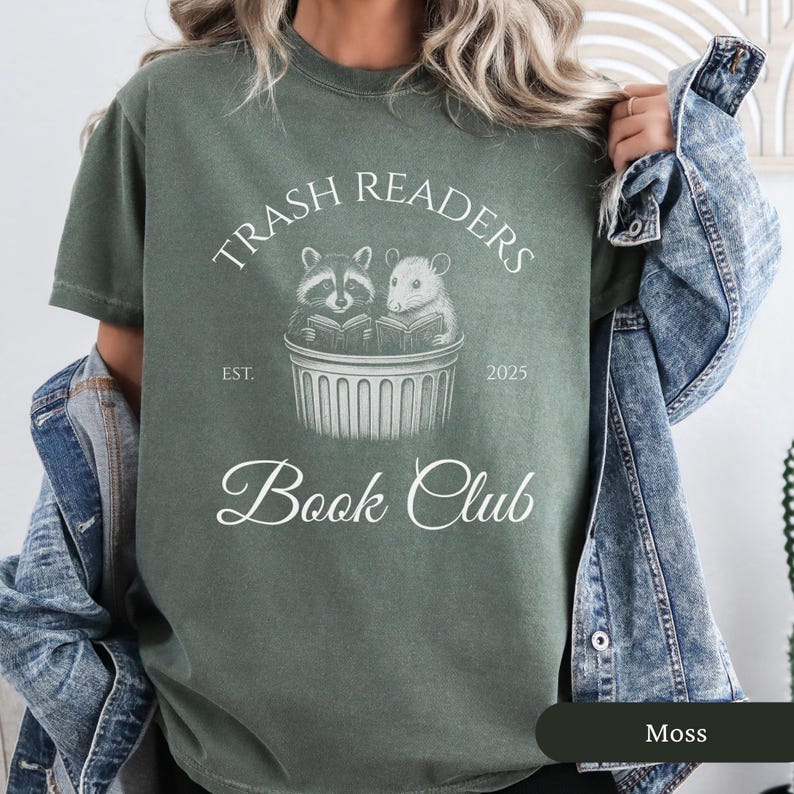 Puede incluir: Una camiseta verde con un dise&ntilde;o gr&aacute;fico blanco que dice "Trash Readers Book Club" con un mapache y una rata leyendo libros en un contenedor de basura. El texto tambi&eacute;n dice "Est. 2025".