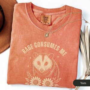 May include: A coral-coloured Comfort Colors t-shirt featuring the text "RAGE CONSUMES ME" above an opossum illustration. The design includes floral elements and stars. A gold necklace with a pendant is draped on the shirt.