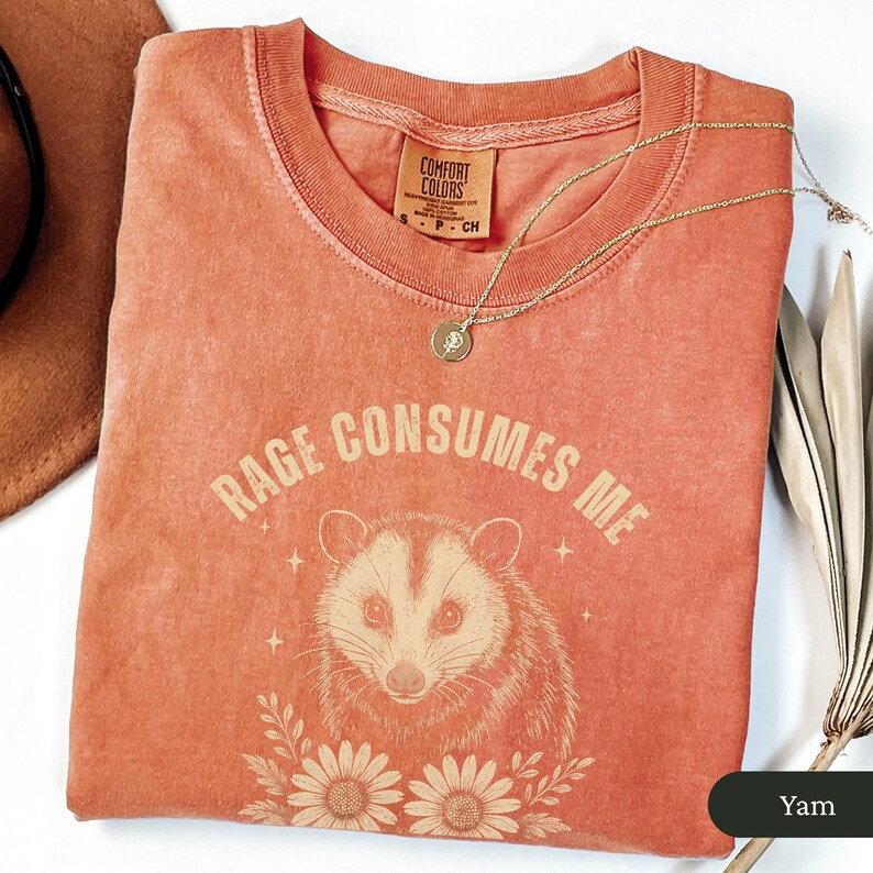 May include: A coral-coloured Comfort Colors t-shirt featuring the text "RAGE CONSUMES ME" above an opossum illustration. The design includes floral elements and stars. A gold necklace with a pendant is draped on the shirt.