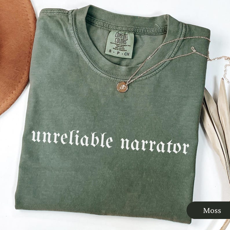 May include: A moss green t-shirt with the phrase "unreliable narrator" in white gothic font. A gold necklace with a pendant rests on the shirt. The shirt has a crew neck and a tag that reads "Comfort Colors".