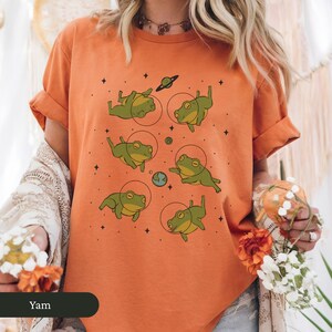 May include: Orange t-shirt featuring a whimsical design of green frogs in space suits, surrounded by planets and stars. The shirt has short sleeves and a relaxed fit, perfect for casual wear. The word "Yam" is visible at the bottom.