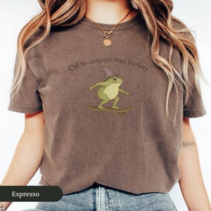 Puede incluir: Camiseta color espresso con una rana de dibujos animados con un sombrero de mago y montada en monopat&iacute;n. El texto "Off to commit tomfoolery" est&aacute; encima de la rana. La camiseta es de cuello redondo y manga corta.