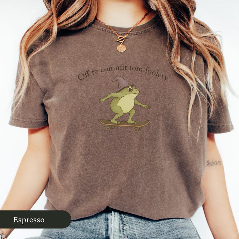 Puede incluir: Camiseta color espresso con una rana de dibujos animados con un sombrero de mago y montada en monopat&iacute;n. El texto "Off to commit tomfoolery" est&aacute; encima de la rana. La camiseta es de cuello redondo y manga corta.