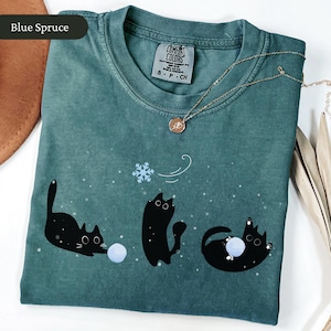 Puede incluir: Camiseta Comfort Colors azul abeto con un dise&ntilde;o l&uacute;dico de tres gatos negros interactuando con un copo de nieve y bolas azules. La camiseta tiene cuello redondo y mangas cortas. Un collar dorado con un colgante est&aacute; sobre la camiseta.
