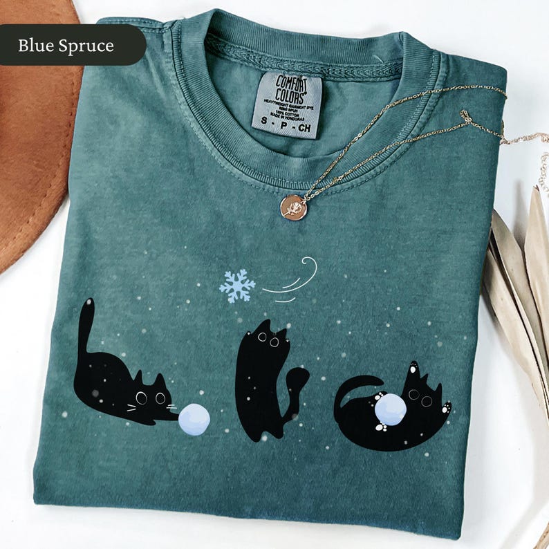Puede incluir: Camiseta Comfort Colors azul abeto con un dise&ntilde;o l&uacute;dico de tres gatos negros interactuando con un copo de nieve y bolas azules. La camiseta tiene cuello redondo y mangas cortas. Un collar dorado con un colgante est&aacute; sobre la camiseta.
