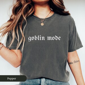 May include: A dark gray t-shirt with the words "goblin mode" printed in white gothic lettering. The shirt is a solid color and has a relaxed fit. The word "Pepper" is in a dark green rectangle at the bottom.
