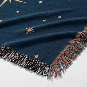 Puede incluir: Una manta azul marino con un dise&ntilde;o celestial. La manta presenta estrellas doradas y un borde de flecos con hilos negros, blancos y multicolores. La manta est&aacute; doblada, mostrando el patr&oacute;n y los flecos.
