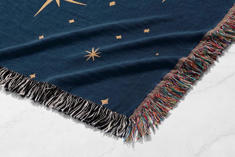 Puede incluir: Una manta azul marino con un dise&ntilde;o celestial. La manta presenta estrellas doradas y un borde de flecos con hilos negros, blancos y multicolores. La manta est&aacute; doblada, mostrando el patr&oacute;n y los flecos.