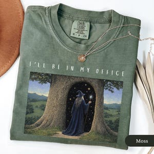 May include: Moss green t-shirt with the text "I'll be in my office." The graphic depicts a wizard in a blue robe standing in a tree doorway. A gold necklace with a pendant is draped on the shirt.