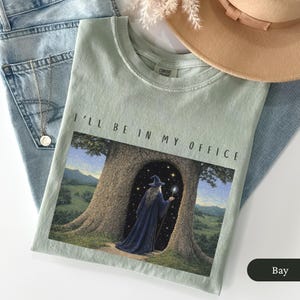 Op de afbeelding: Lichtgroen t-shirt met de tekst "I'll Be In My Office" boven een afbeelding van een tovenaar in een blauwe jurk die in een boomdeur staat. Het shirt is op een blauwe jeans gevouwen.