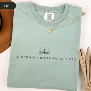 May include: A light green Comfort Colors t-shirt with the text "I CLOSED MY BOOK TO BE HERE" and an illustration of an open book. A gold necklace with a pendant rests on the shirt.