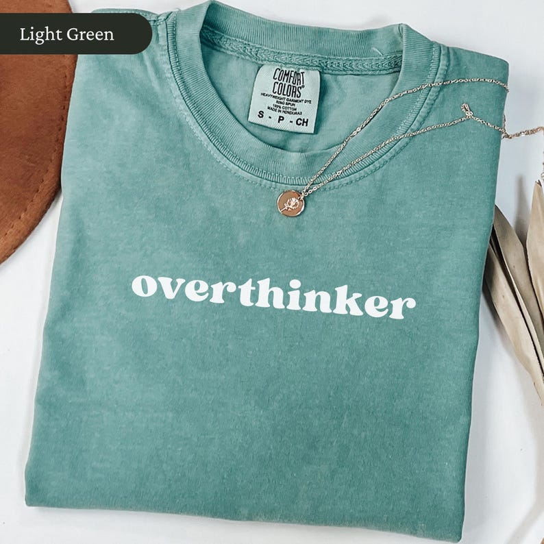 May include: Light green t-shirt with the word "overthinker" in white block letters. A silver necklace with a small pendant rests on the shirt. The shirt is folded and has a tag that reads "Comfort Colors".