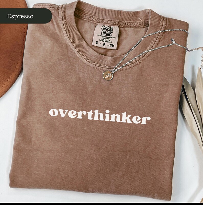 May include: Espresso-coloured t-shirt with the word "overthinker" in white block letters. A silver necklace with a small pendant rests on the shirt. The shirt has a Comfort Colors tag.