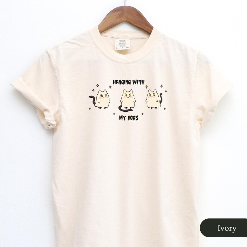 May include: Ivory t-shirt featuring the text "Hanging with My Boos" and a graphic of three ghost cats. The cats have yellow eyes and black tails. The design is centered on the front of the shirt.