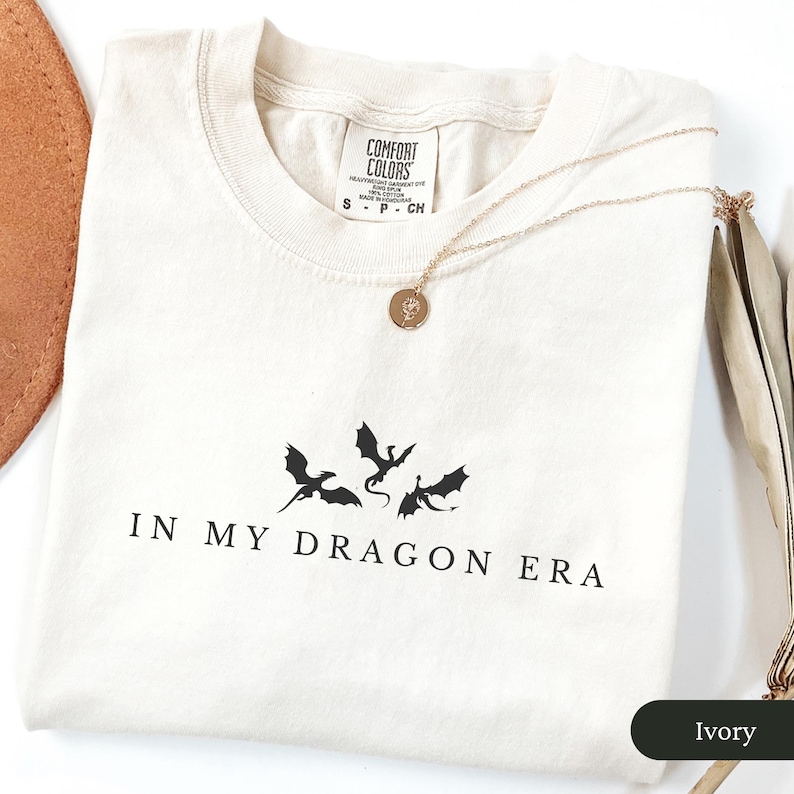 May include: Ivory-colored t-shirt with the phrase "IN MY DRAGON ERA" and a graphic of three dragons in black. A gold necklace with a round pendant rests on the shirt. The shirt is made by Comfort Colors.