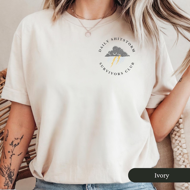 May include: Ivory-colored t-shirt with a round graphic featuring a cloud, lightning bolts, and the text "DAILY SHITSTORM SURVIVORS CLUB." The shirt has short sleeves and a crew neck. The color of the shirt is ivory.