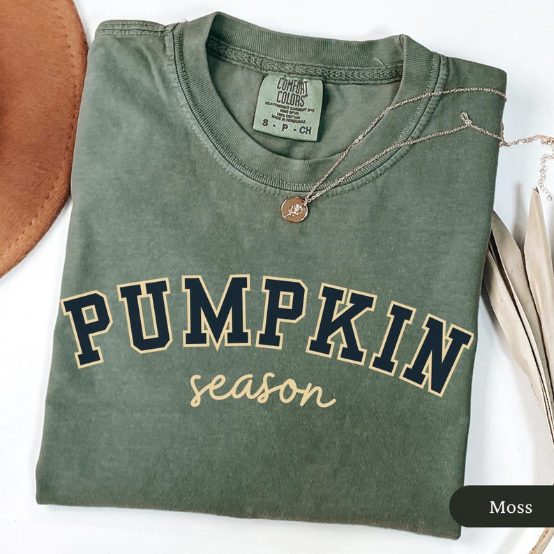 Pu&ograve; includere: Una maglietta verde muschio con le parole "PUMPKIN season" stampate sul davanti in blu navy e beige. Una collana d'oro con un ciondolo &egrave; drappeggiata sulla maglietta. La maglietta &egrave; piegata e presentata su una superficie bianca.