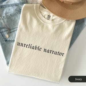 May include: Ivory-colored t-shirt with the phrase "unreliable narrator" printed in black gothic font. The shirt is folded and rests on a pair of blue jeans. A tan hat is partially visible in the background.