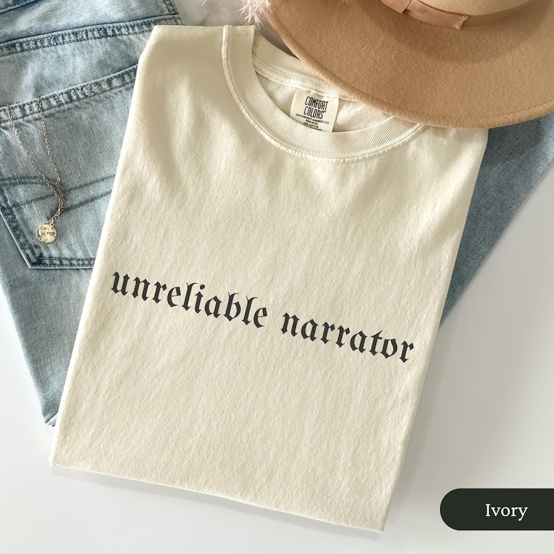 May include: Ivory-colored t-shirt with the phrase "unreliable narrator" printed in black gothic font. The shirt is folded and rests on a pair of blue jeans. A tan hat is partially visible in the background.