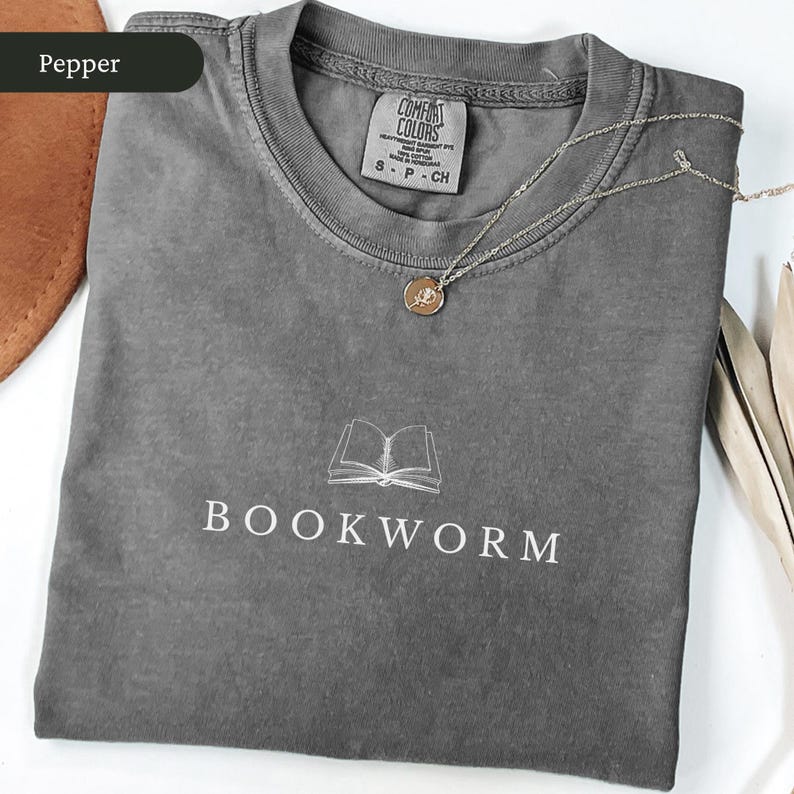 Op de afbeelding: Een grijze t-shirt met het woord "BOOKWORM" in het wit gedrukt onder een lijntekening van een open boek. Een goudkleurige ketting met een kleine hanger ligt op de shirt. De shirt is opgevouwen en heeft een label met "Comfort Colors".
