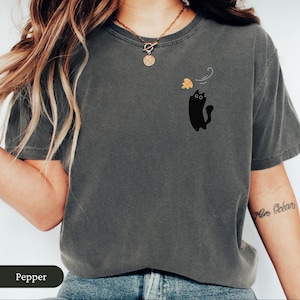 May include: A dark grey t-shirt featuring a black cat graphic and a falling leaf design. The cat has large eyes and a curved tail. The word "Pepper" is at the bottom of the image.