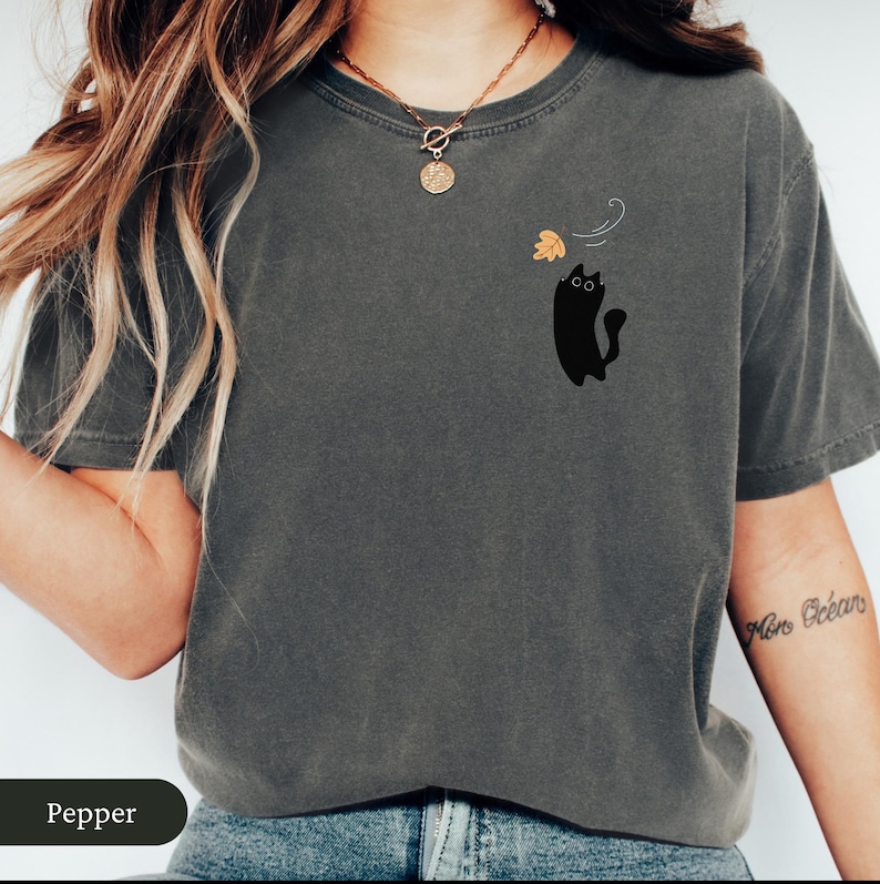May include: A dark grey t-shirt featuring a black cat graphic and a falling leaf design. The cat has large eyes and a curved tail. The word "Pepper" is at the bottom of the image.