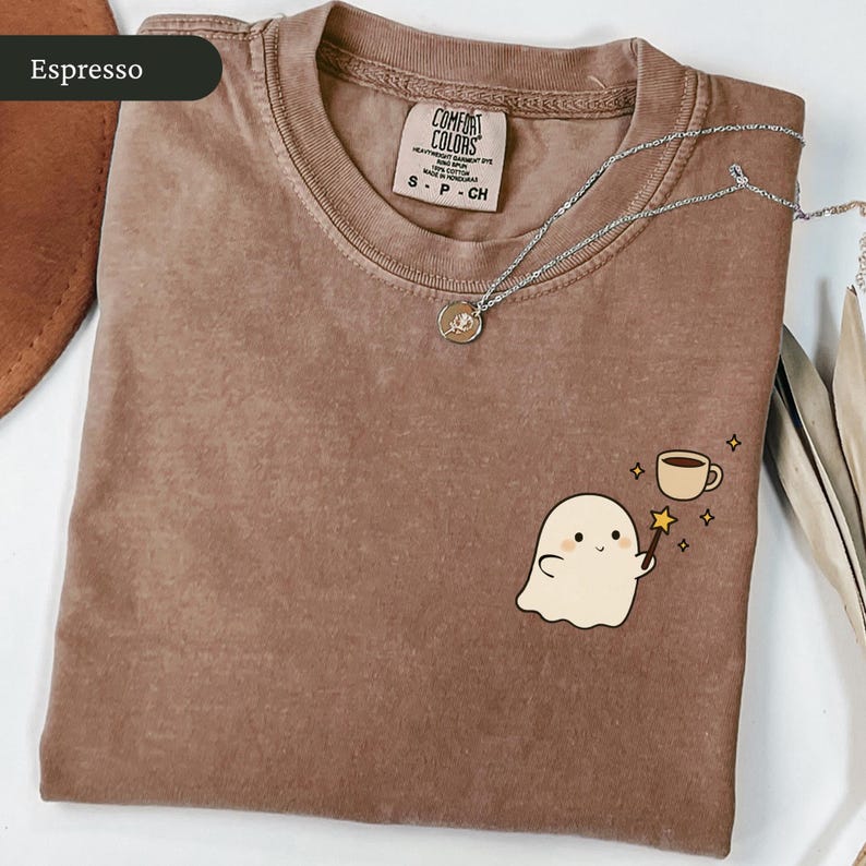 May include: Espresso-colored Comfort Colors t-shirt with a ghost graphic holding a magic wand and a coffee cup. A silver necklace with a small pendant rests on the shirt. The shirt is size S.